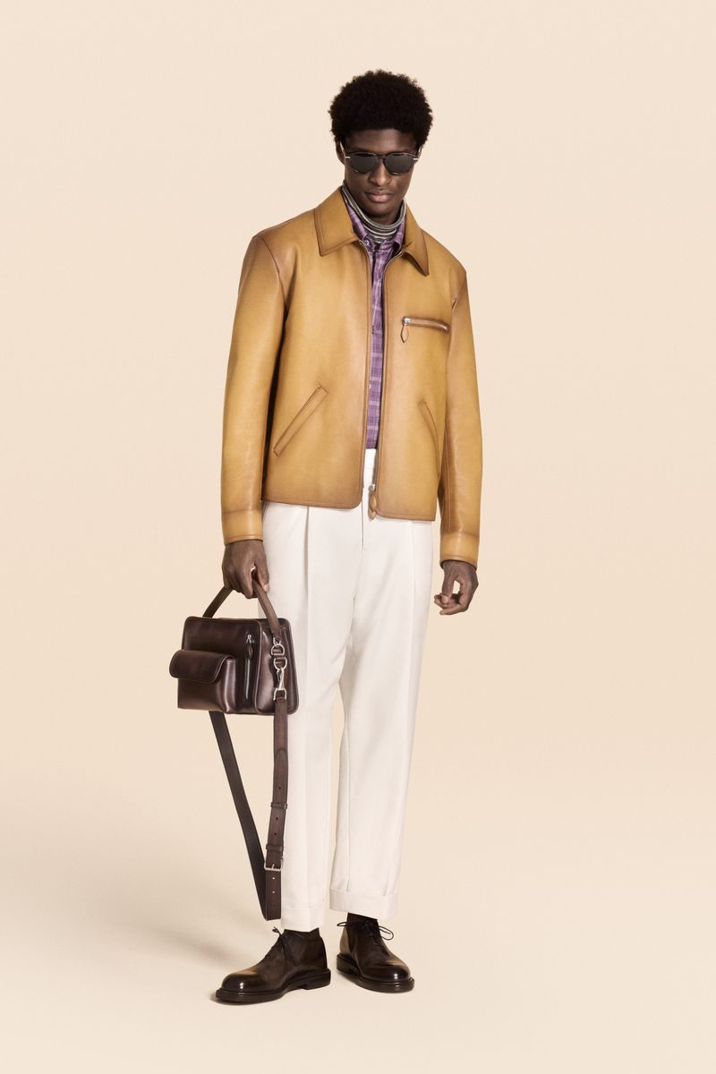 A stylish Black male model stands in a studio wearing a tan jacket, white trousers, black shoes, and sunglasses, holding a dark leather bag.