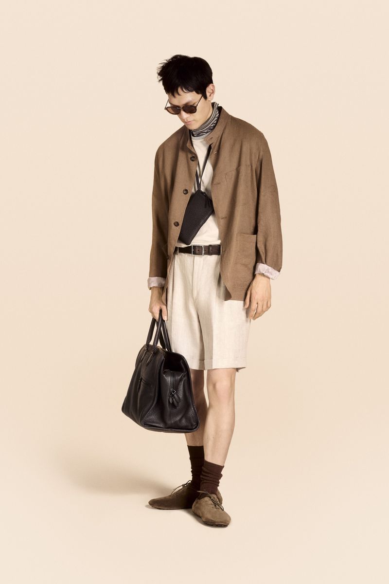 Man stands wearing brown jacket, light shorts, and sunglasses, holding a black leather duffel bag against a beige background.