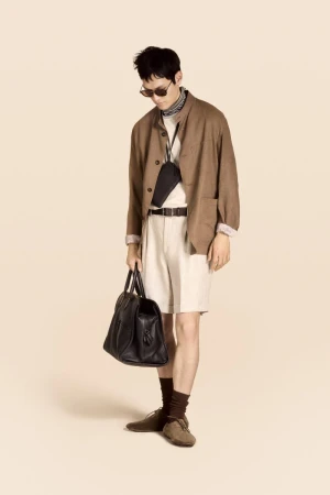 Man stands wearing brown jacket, light shorts, and sunglasses, holding a black leather duffel bag against a beige background.
