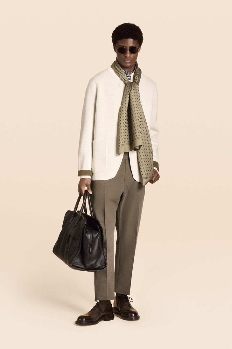 A stylish man in a cream jacket, brown pants, and sunglasses holds a black leather bag, standing in front of a neutral background.