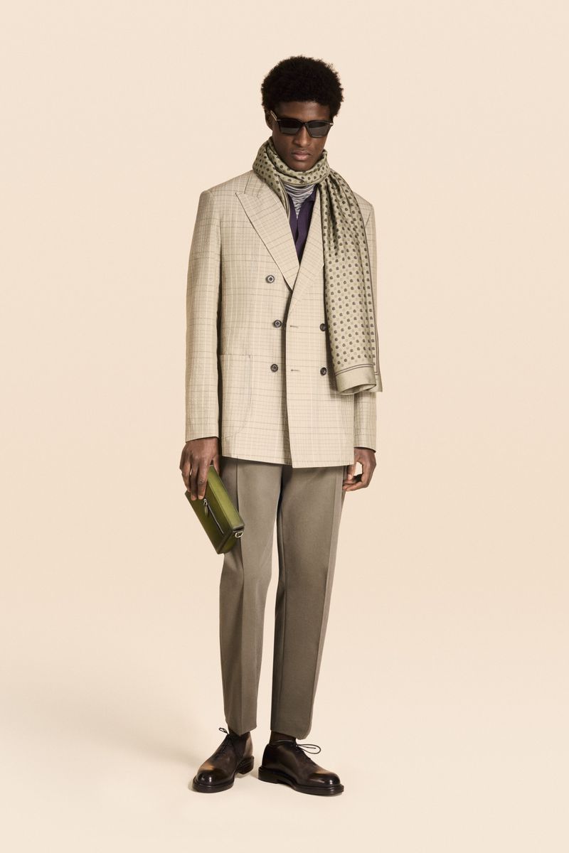 An African American man in a beige blazer, olive pants, and a patterned scarf poses against a neutral backdrop, holding a green clutch.