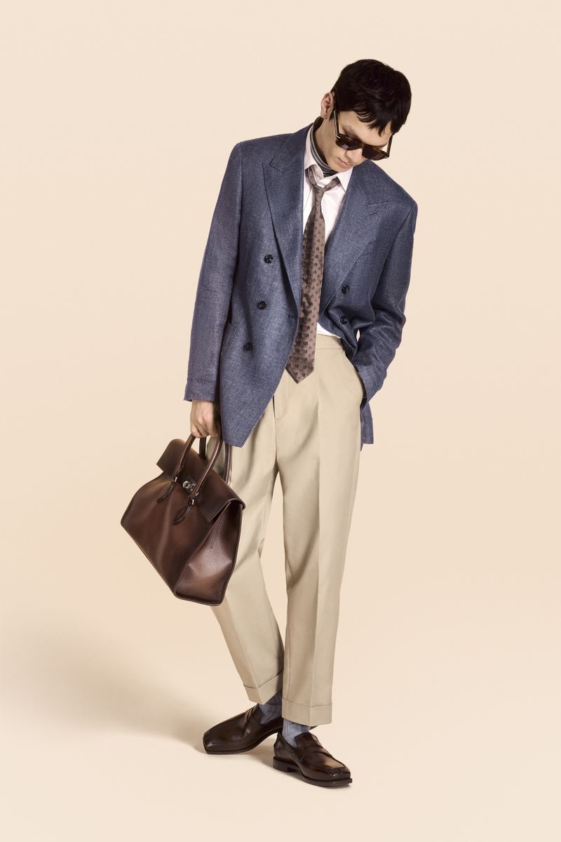 Man in blue blazer, tie, and tan pants carries a brown leather bag in a full shot against a beige background; studio lighting, fashion photography.