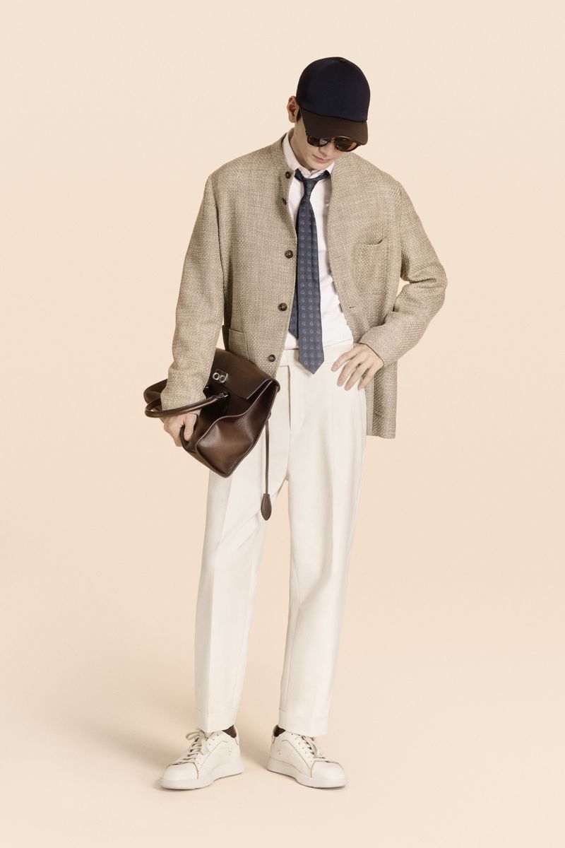 Full shot of adult male in neutral tones with a hat and sunglasses, wearing a blazer, tie, and white trousers, carrying a leather bag.