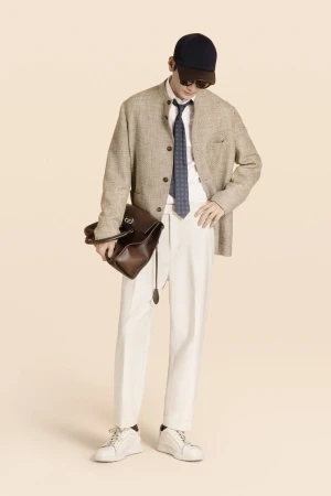 Full shot of adult male in neutral tones with a hat and sunglasses, wearing a blazer, tie, and white trousers, carrying a leather bag.
