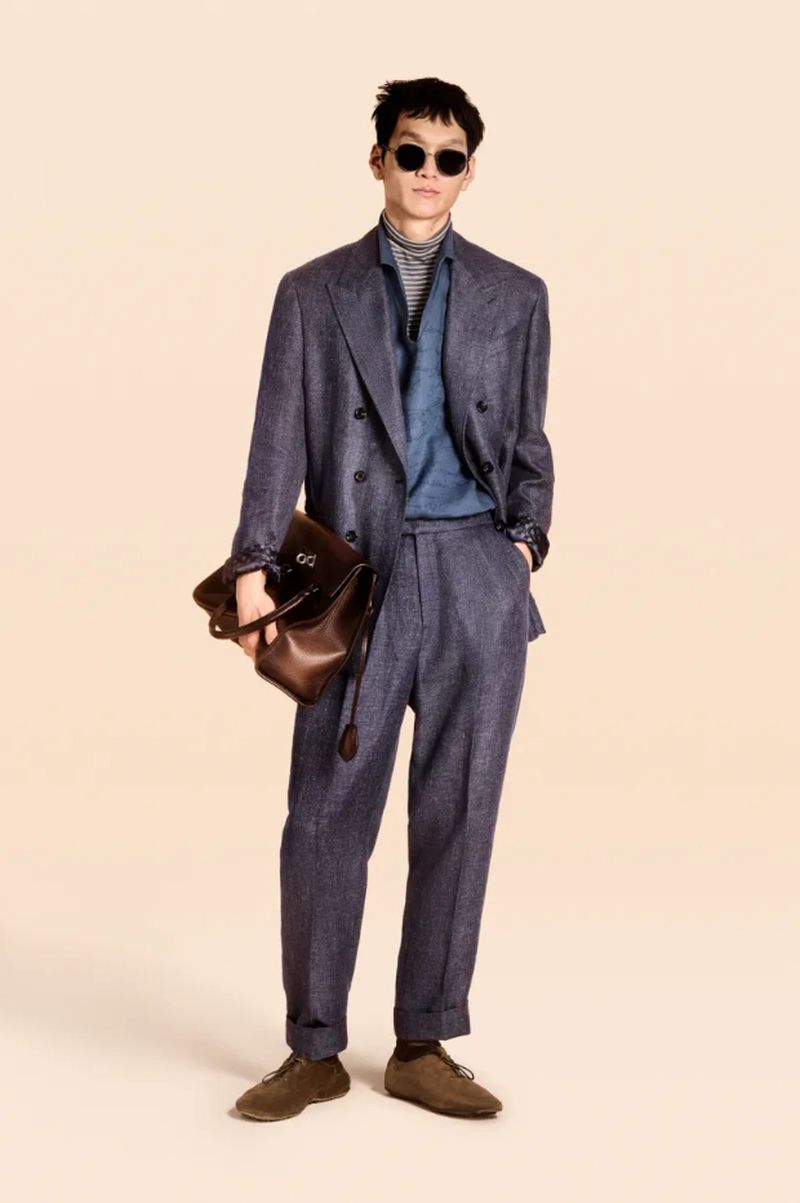 Man in a blue suit poses with a brown leather bag and sunglasses in a studio. The neutral backdrop highlights the fashion-forward look.