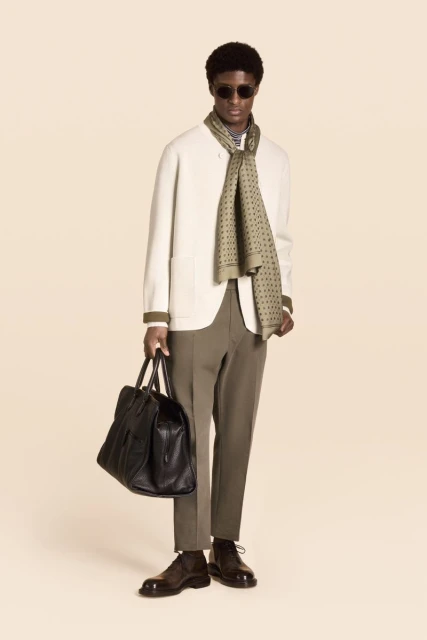 Model in off-white jacket, green patterned scarf and holding a black leather bag against a neutral studio backdrop