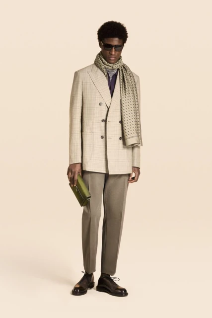 Model wearing a plaid double-breasted blazer, patterned scarf, and holding a clutch against a warm neutral background.