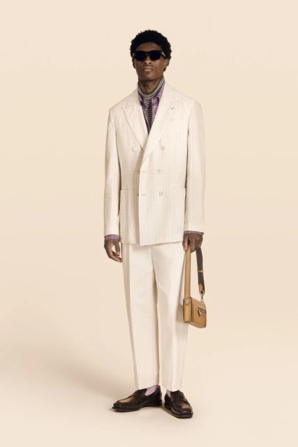Model Aboubakar Konte standing in a cream double-breasted suit, sunglasses, and carrying a tan leather bag on a cream backdrop.