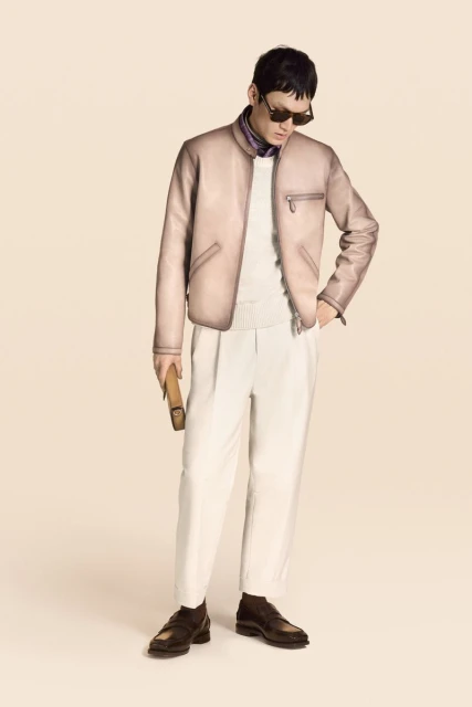 East Asian male model wearing a pale leather bomber jacket, sunglasses, and off-white pleated trousers against a warm studio background.