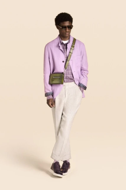 Model wearing a lilac blazer, light trousers, sunglasses, and an olive green crossbody bag against a warm studio backdrop.