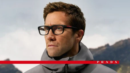 Jake Gyllenhaal wearing black rectangular Prada Linea Rossa optical glasses and a grey technical jacket with mountains in the background.