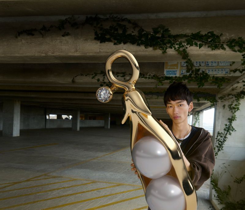 Model Bai holds an oversized gold pea pod charm with large pearl peas in an open concrete carpark.