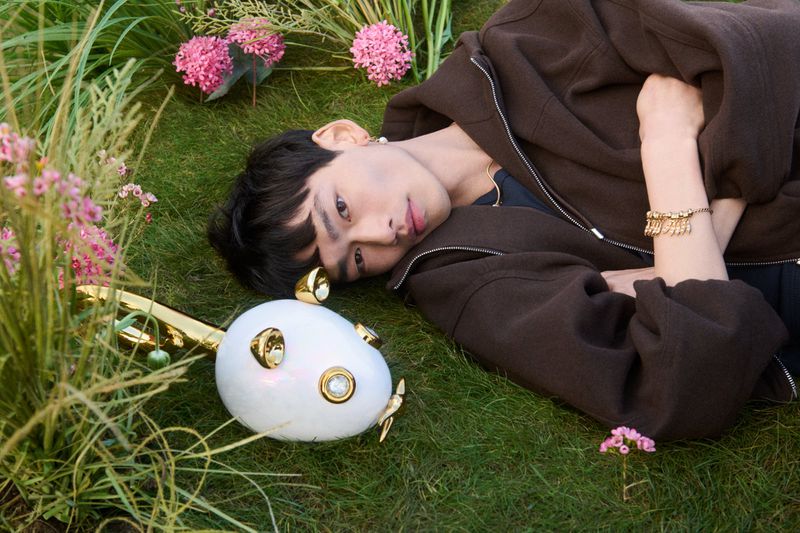 Model Bai lying on a grass lawn next to pink flowers and a white and gold egg-shaped sculpture for Pandora.