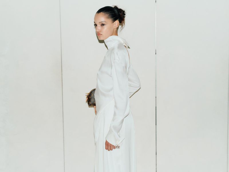 Model Corinne Murray wearing a high-neck white silk dress and holding a monogram pouch against a white wall.