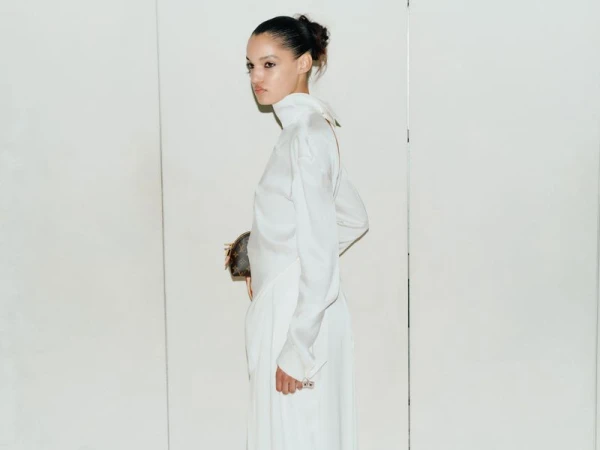 Model Corinne Murray wearing a high-neck white silk dress and holding a monogram pouch against a white wall.