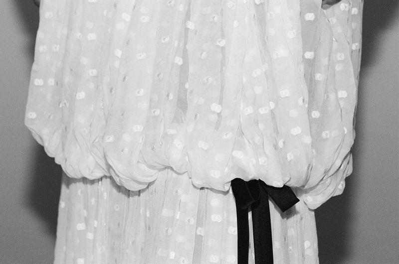 Close-up detail of a white textured dotted fabric dress with a gathered blouson waist and black ribbon tie in a studio setting.