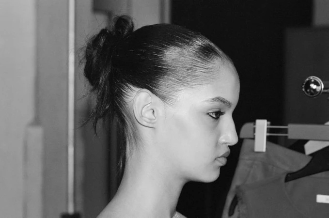 Black and white profile portrait of model Corinne Murray with a slicked-back bun backstage by clothing racks.