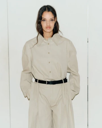 Model Corinne Murray wearing an oversized beige button-up shirt and wide-leg trousers with a black belt against a neutral backdrop.