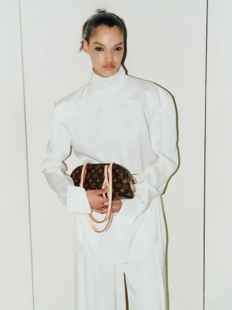 Model Corinne Murray wearing an all-white high-neck tunic outfit and holding a monogram canvas handbag against a pale backdrop.