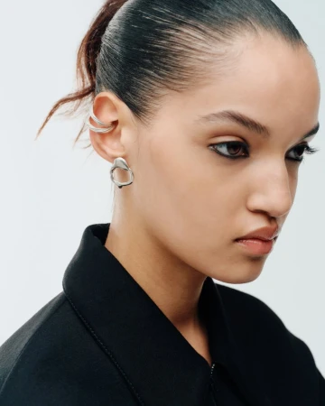 Close-up portrait of model Corinne Murray wearing silver sculpted earrings and a black jacket against a white studio background.