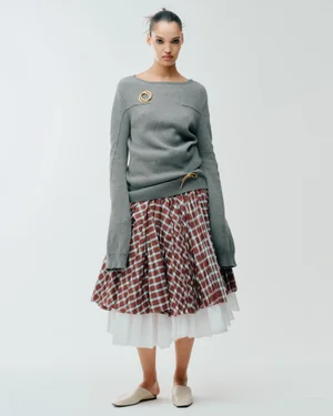 Model Corinne Murray posing in a long-sleeve grey sweater and layered brown plaid skirt on a white background.