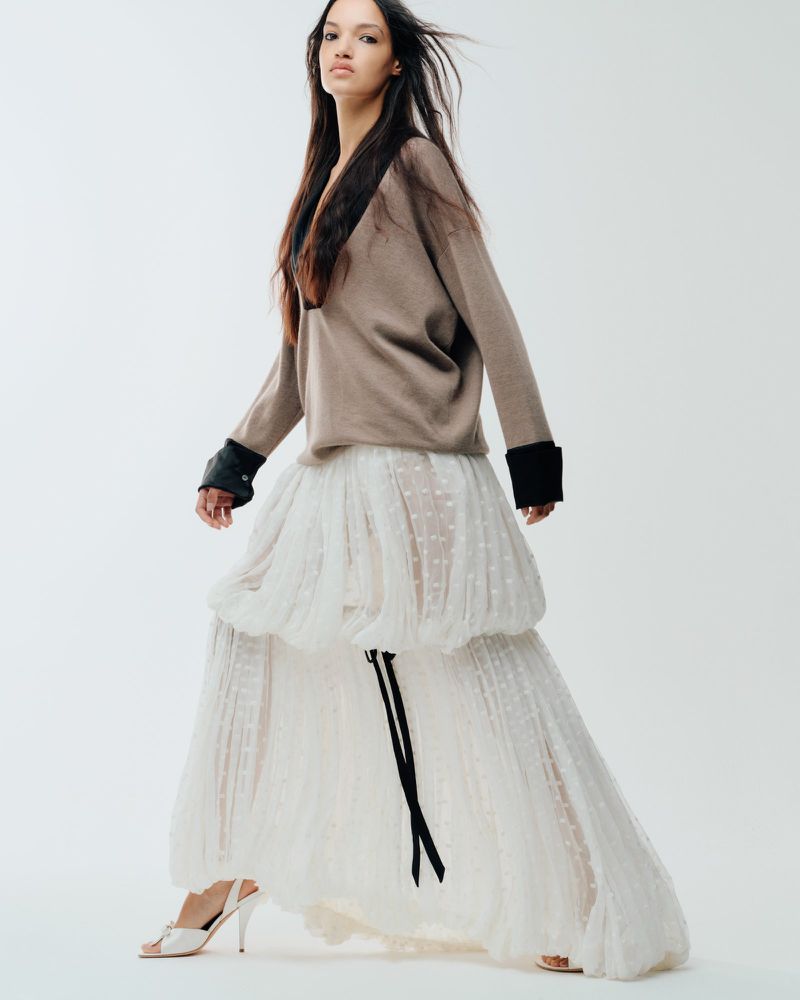 Model Corinne Murray posing in a taupe sweater and voluminous tiered white maxi skirt against a clean white seamless backdrop.