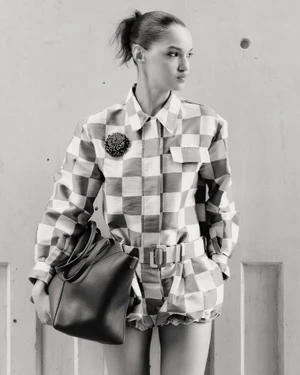 Model Corinne Murray wearing a checkered belted romper and holding a large leather tote bag against a concrete wall in black and white.