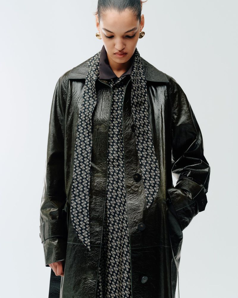 Corinne Murray looking down wearing a glossy black leather coat and patterned long scarf against a white studio background.