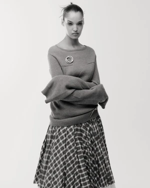 Model Corinne Murray in a knit sweater with a circular brooch and pleated plaid skirt against a plain studio background.