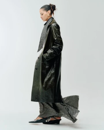 Corinne Murray posing in profile wearing a glossy dark trench coat over a patterned floor-length dress against a white studio backdrop.