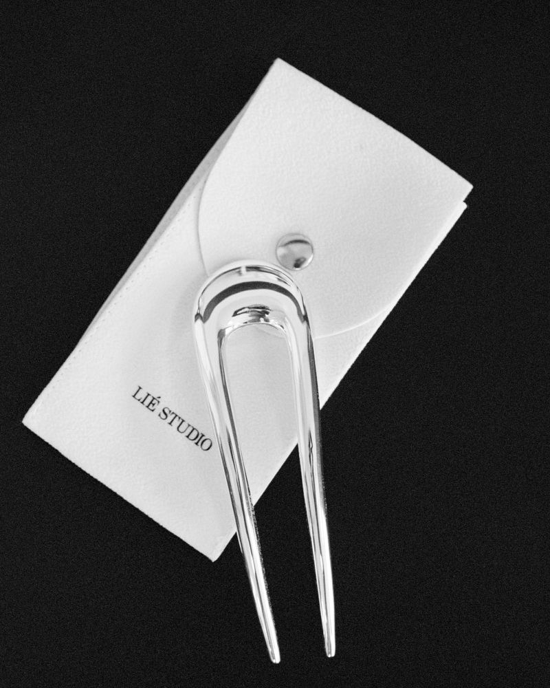Polished silver metallic hair pin resting on a white leather snap pouch over a textured black background.