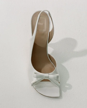 Top-down view of a white satin slingback high-heeled shoe with a front bow detail on an off-white background.