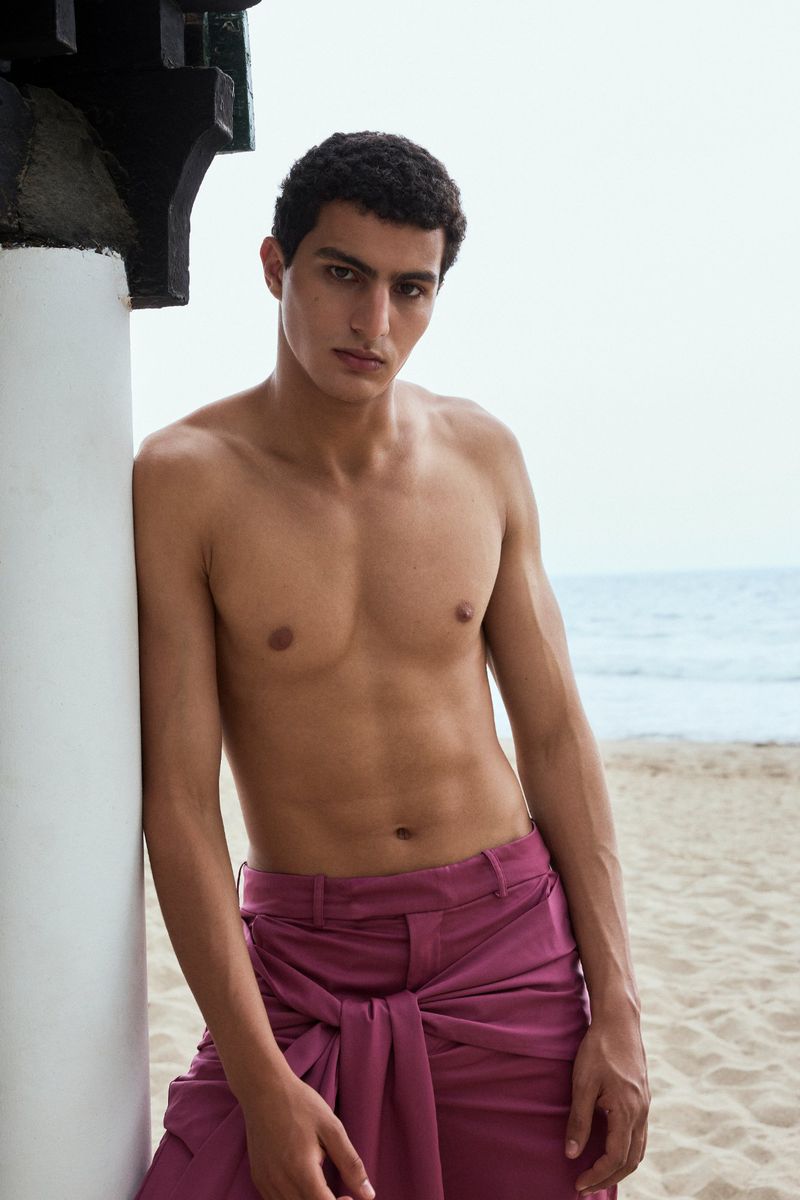 Shirtless man with short curly hair leaning against a white pillar on a sandy beach wearing plum-pink tailored trousers.