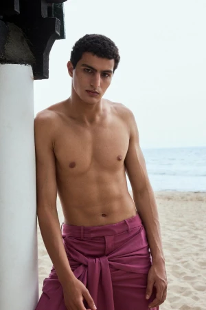 Shirtless man with short curly hair leaning against a white pillar on a sandy beach wearing plum-pink tailored trousers.