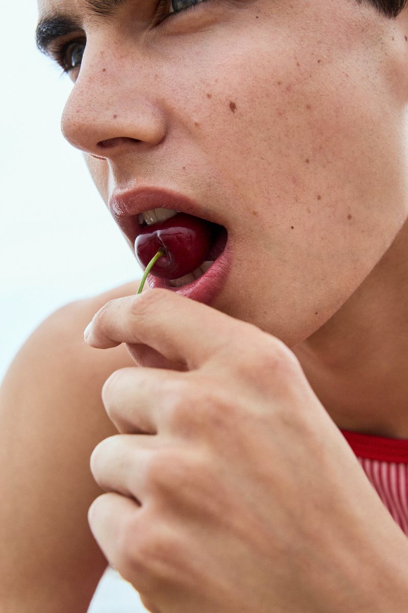 Close-up portrait of a young man eating a fresh dark red cherry held between his lips against a bright open background.