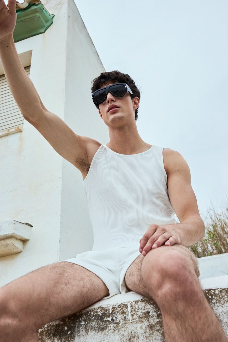 Low-angle shot of a young man in black sunglasses and a white tank top seated outdoors on a whitewashed ledge.