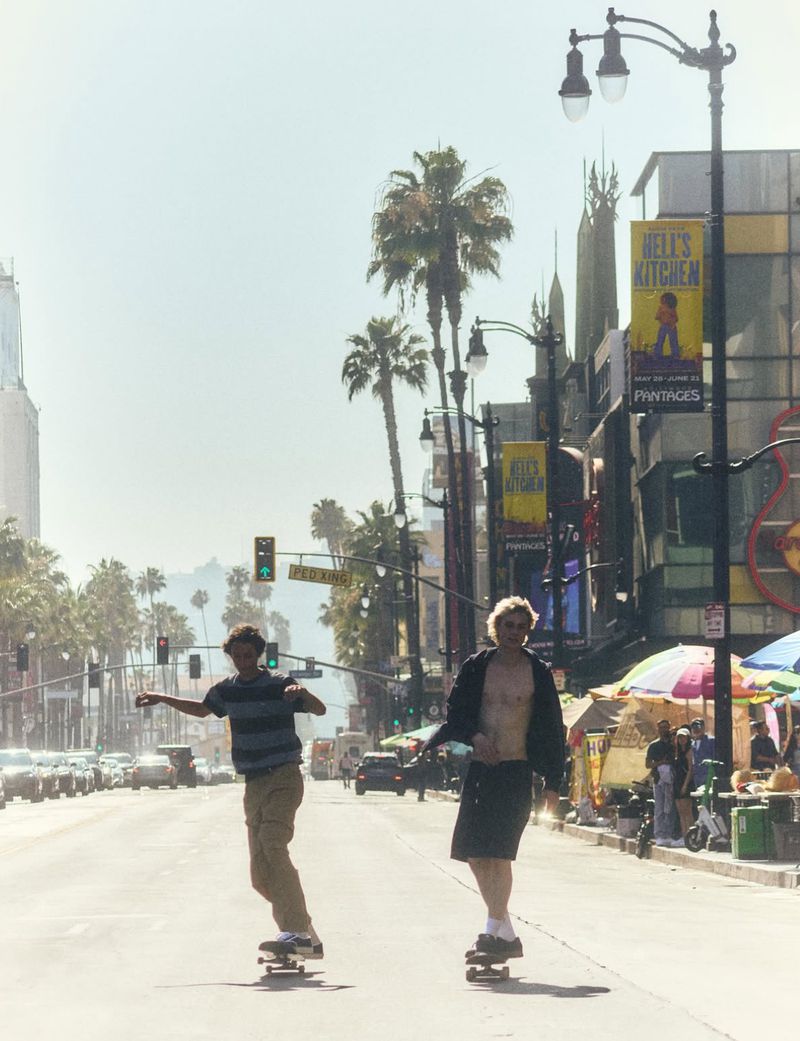 Two young men skate down a wide city boulevard lined with palm trees, streetlights, and theater banners in bright daylight.