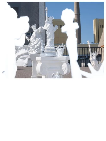 Collection of white Statue of Liberty sculptures and busts arranged on an outdoor terrace with industrial architecture behind.