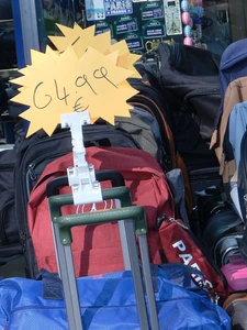 Luggage, backpacks, and a blue duffel bag displayed with a yellow starburst price tag reading 64.99 euros at a Paris souvenir stall.