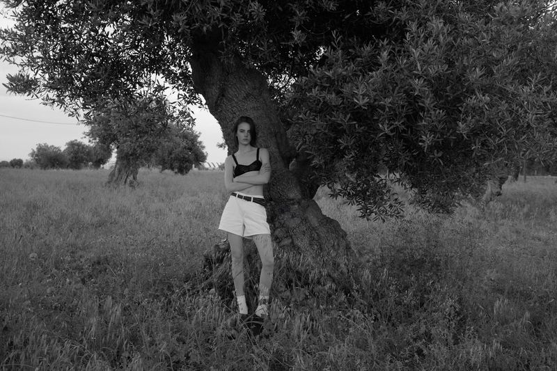 Monochrome shot of a woman in a black bralette and white shorts standing beneath a large tree in a grassy field.
