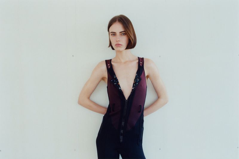 Model Karyna Maziar wearing a deep V-neck sleeveless jumpsuit against a plain white studio background.