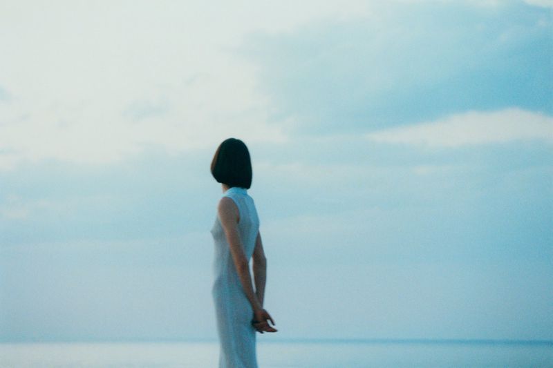 Model Karyna Maziar wearing a pale sleeveless dress standing in profile before the sea for Vogue Polska.