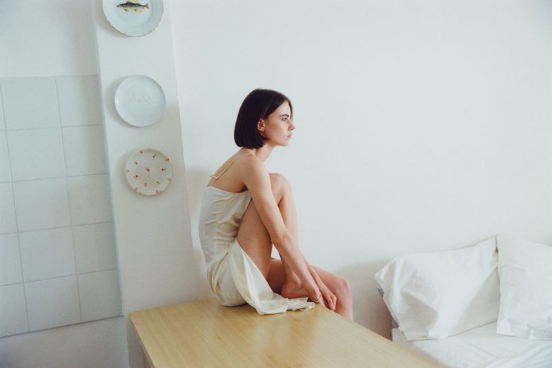 Model Karyna Maziar wearing a cream slip dress seated with knees tucked on a wooden surface in a white bedroom.
