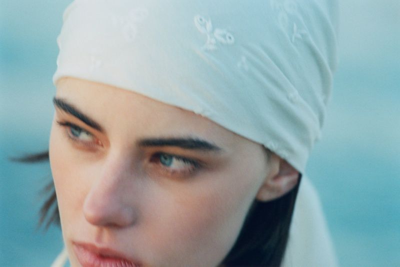 Close-up portrait of model Karyna Maziar wearing a white embroidered headscarf against a soft blue background for Vogue Polska.