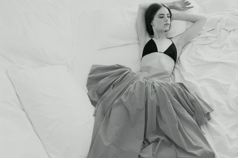 Model Karyna Maziar reclining on a white bed wearing a black triangle top and a voluminous skirt for Vogue Polska.