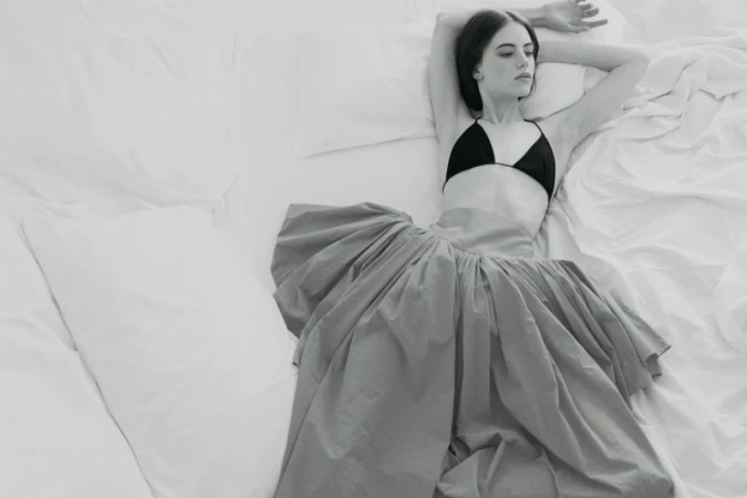 Model Karyna Maziar reclining on a white bed wearing a black triangle top and a voluminous skirt for Vogue Polska.