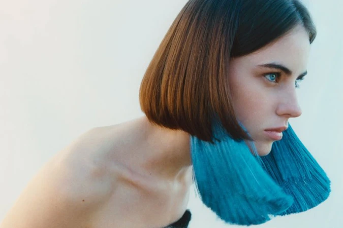 Close-up portrait of a model with a sleek brown bob and oversized bright teal tassel earrings against a pale seamless background.