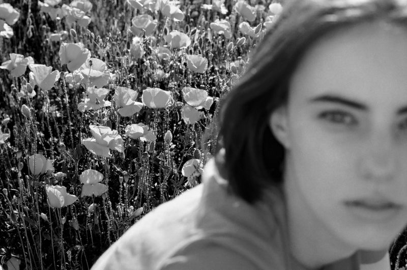 Black and white portrait of a woman in soft focus in the foreground, with a field of blooming poppies in the background.