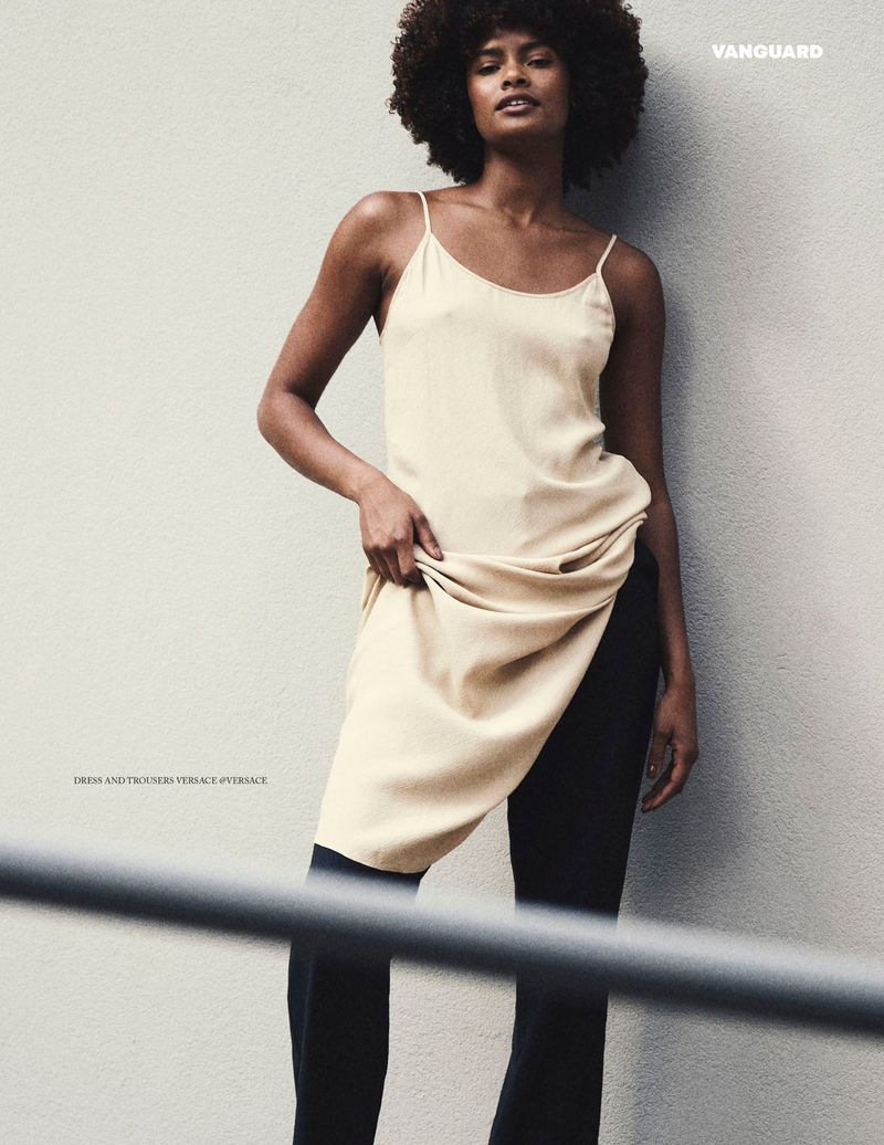A woman with an afro wearing a cream spaghetti-strap slip dress over black tailored trousers against a textured neutral wall.
