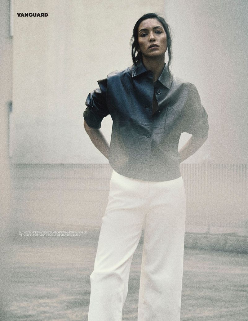 Fashion model wearing a dark leather button-up shirt and wide-leg white trousers in a hazy outdoor setting.